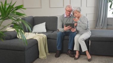 Tabletli yaşlı çift internetten video izliyor. Karı koca evde vakit geçiriyor. Mutlu modern olgun yaşlı çift aile eğlencesi için tablet kullanıyor. Yaşlılar modern teknoloji