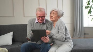 Tabletli yaşlı çift internetten video izliyor. Karı koca evde vakit geçiriyor. Mutlu modern olgun yaşlı çift aile eğlencesi için tablet kullanıyor. Yaşlılar modern teknoloji