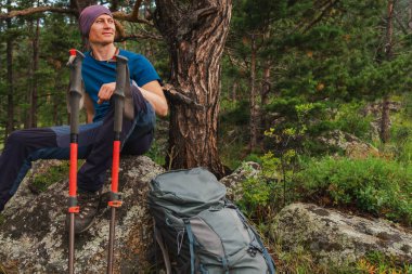 Uçuruma tırmanma macerası. Sırt çantalı adam güzel manzaraya bakıyor. Sırt çantalı gezgin yeşil orman ve gölün üzerindeki büyük kayanın üzerinde oturuyor. Gülümseyen mutlu yürüyüşçü, yürüyüş turizminin keyfini çıkar.