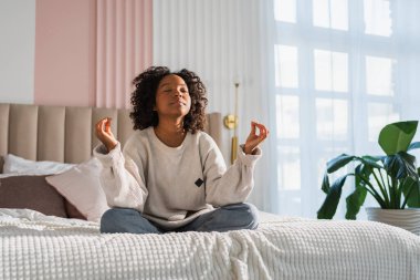 Yoga farkındalığı meditasyonu. Genç sağlıklı Afrikalı kız evde yoga yapıyor. Nilüfer çiçekli bir kadın yatakta oturmuş, meditasyon yapıyor, gülümsüyor, evde dinleniyor. Nefes egzersizi yapan bir kız. Evde yoga.