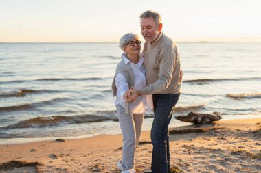 Yürümeye devam. Romantik olgun çift plaj gezisinde birlikte dans ediyor. Mutlu gülümseyen aile emeklisi kadın koca eş birlikte eğleniyor. Aile zamanı aşk bakımı