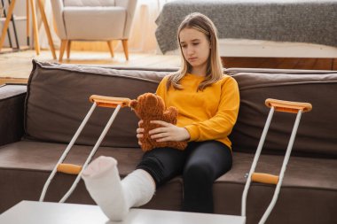 Kanepede oturan kırık bacaklı genç kız. Üzgün küçük çocuk, kırık bacağını iyileştirmek için oyuncak ayısına sarılıyor. Alçı ve koltuk değneği kullanan bir kız. Kaza sonucu oluşan kırık, ayak bandajı.