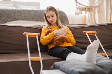 Kanepede oturan kırık bacaklı genç kız. Üzgün küçük çocuk, kırık bacağını iyileştirmek için oyuncak ayısına sarılıyor. Alçı ve koltuk değneği kullanan bir kız. Kaza sonucu oluşan kırık, ayak bandajı.