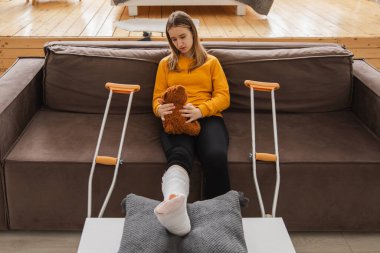 Kanepede oturan kırık bacaklı genç kız. Üzgün küçük çocuk, kırık bacağını iyileştirmek için oyuncak ayısına sarılıyor. Alçı ve koltuk değneği kullanan bir kız. Kaza sonucu oluşan kırık, ayak bandajı.