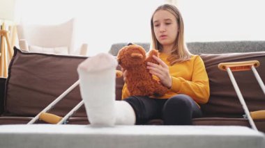 Kanepede oturan kırık bacaklı genç kız. Üzgün küçük çocuk, kırık bacağını iyileştirmek için oyuncak ayısına sarılıyor. Alçı ve koltuk değneği kullanan bir kız. Kaza sonucu oluşan kırık, ayak bandajı.