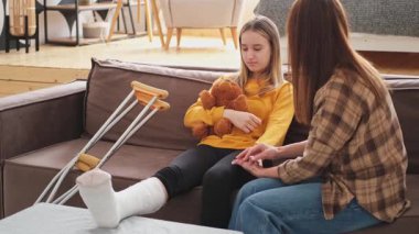 Kırık bacağı alçıda olan bir kız annesiyle evde kanepede oturuyor. Kırık bacağı iyileşen üzgün küçük çocuk oyuncak ayıya sarılıyor. Hızlı rehabilitasyon için koltuk değneklerini kullanan bir kız. Anne destek kızı
