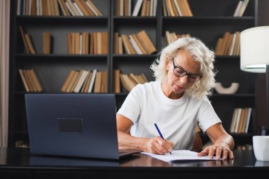 İş yerinde dizüstü bilgisayar kullanarak not yazan orta yaşlı bir kadın. Yetişkin, yaşlı bir iş kadınının ofiste çalışıp, online işler yaparak yazılı plaklar çıkarmasına odaklanmış. Olgun patron profesyonel işçi.