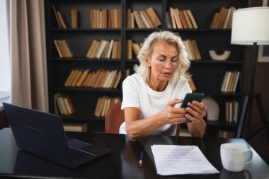 İş yerinde akıllı telefon kullanan kendine güvenen, Avrupalı orta yaşlı bir kadın. Ofisinde cep telefonu olan 60 'lı yaşlarda şık, olgun bir iş kadını. Patron internet uygulamaları kullanıyor
