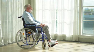 Tekerlekli sandalyede oturan üzgün yaşlı adam huzurevinin penceresinden dışarı bakıyor. Engelli insanlar için tekerlekli sandalyedeki yaşlı felçli adam depresyonda. Yalnız yaşlı hasta adam tekerlekli sandalyede oturuyor.