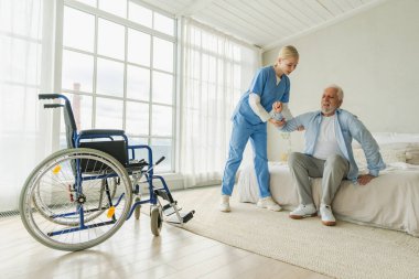 Rehabilitasyon sağlık hizmetleri. Fizyoterapist hemşire tekerlekli sandalyedeki yaşlı adamın ayağa kalkmasına yardım ediyor. Engelli insanlar için sandalyedeki adam hemşire yardımı ile birkaç adım atıyor. Tıbbi bakıcı.