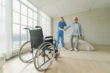 Rehabilitasyon sağlık hizmetleri. Fizyoterapist hemşire tekerlekli sandalyedeki yaşlı adamın ayağa kalkmasına yardım ediyor. Engelli insanlar için sandalyedeki adam hemşire yardımı ile birkaç adım atıyor. Tıbbi bakıcı.