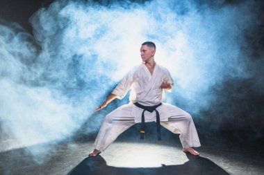 Siyah kuşaklı karate dövüş sanatları için beyaz kimonolu güçlü adam siyah arka plandaki dövüş eğitimine hazırlanıyor. Antrenman hareketleri yumruklar gölge dövüşü alıştırmaları antrenmana hazır.