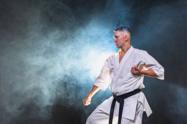 Siyah kuşaklı karate dövüş sanatları için beyaz kimonolu güçlü adam siyah arka plandaki dövüş eğitimine hazırlanıyor. Antrenman hareketleri yumruklar gölge dövüşü alıştırmaları antrenmana hazır.