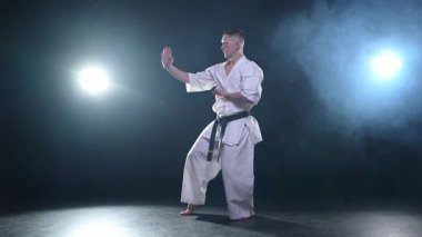 Siyah kuşaklı karate dövüş sanatları için beyaz kimonolu güçlü adam siyah arka plandaki dövüş eğitimine hazırlanıyor. Antrenman hareketleri yumruklar gölge dövüşü alıştırmaları antrenmana hazır.