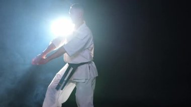Siyah kuşaklı karate dövüş sanatları için beyaz kimonolu güçlü adam siyah arka plandaki dövüş eğitimine hazırlanıyor. Antrenman hareketleri yumruklar gölge dövüşü alıştırmaları antrenmana hazır.