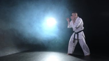 Siyah kuşaklı karate dövüş sanatları için beyaz kimonolu güçlü adam siyah arka plandaki dövüş eğitimine hazırlanıyor. Antrenman hareketleri yumruklar gölge dövüşü alıştırmaları antrenmana hazır.