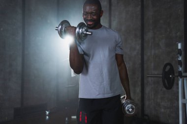 Beden eğitiminde spor. Spor salonunda dambılları toplayan Afrikalı Amerikalı bir vücut geliştirici. Fitness kulübünde halterli kasları çalıştırmak için ağırlık çalışıyorum. Spor yapan bir adam. Sağlıklı yaşam tarzı