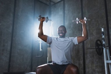 Beden eğitiminde spor. Spor salonunda dambılları toplayan Afrikalı Amerikalı bir vücut geliştirici. Fitness kulübünde halterli kasları çalıştırmak için ağırlık çalışıyorum. Spor yapan bir adam. Sağlıklı yaşam tarzı