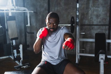 Beden eğitiminde spor. Afrikalı dövüşçü dövüşmeye hazırlanıyor kırmızı boksla yumruk atarken spor sargılarını spor salonunda sarar. Güçlü adam boks antrenmanına hazır yumrukları eğitiyor.