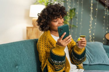 African american woman shopping online holding smartphone paying with gold credit card. Girl sitting at home buying on Internet enter credit card details. Online shopping ecommerce delivery service
