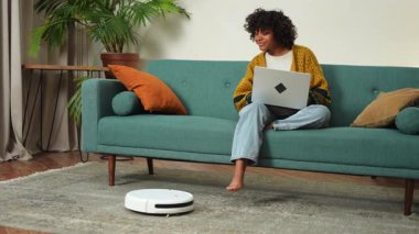 Kanepede dizüstü bilgisayarla çalışan bir kadın için robot süpürge temizleme odası. Dizüstü bilgisayar kullanan bir kadın, yerleri süpüren bir robot. Akıllı ev otomasyonu modern teknoloji günlük hayatta