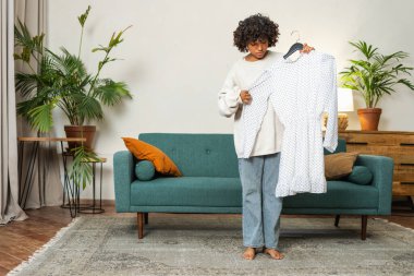 Moda alışverişi. Evde elbise seçen Afrikalı bir kadın. Elbise seçmeye çalışan genç bir kadın. Elbiseleri askıda tutan bir elbise. Kıvırcık esmer tenli, gülümseyen kız elinde askıyla beyaz elbiseyle