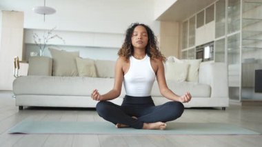 Yoga farkındalığı meditasyonu. Genç sağlıklı Afrikalı kız evde yoga yapıyor. Lotus 'ta oturan kadın yoga minderinde poz veriyor içeride meditasyon yapıyor. Nefes egzersizi yapan bir kız. Evde yoga.