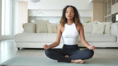 Yoga farkındalığı meditasyonu. Genç sağlıklı Afrikalı kız evde yoga yapıyor. Lotus 'ta oturan kadın yoga minderinde poz veriyor içeride meditasyon yapıyor. Nefes egzersizi yapan bir kız. Evde yoga.