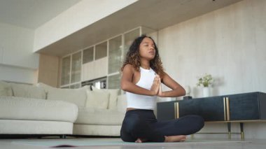 Yoga farkındalığı meditasyonu. Genç sağlıklı Afrikalı kız evde yoga yapıyor. Lotus 'ta oturan kadın yoga minderinde poz veriyor içeride meditasyon yapıyor. Nefes egzersizi yapan bir kız. Evde yoga.