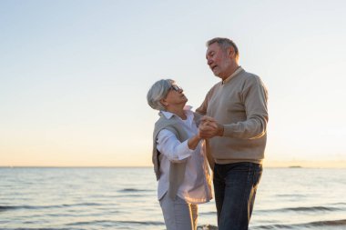Yürümeye devam. Romantik olgun çift plaj gezisinde birlikte dans ediyor. Mutlu gülümseyen aile emeklisi kadın koca eş birlikte eğleniyor. Aile zamanı aşk bakımı