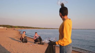 Dünya günü. Gönüllü eylemciler, sahil bölgesindeki çöpleri topluyor. Kadın, okyanus kıyısındaki çöp poşetine plastik şişe koyuyor. Çevre koruma kıyı bölgesi temizliği