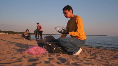Dünya günü. Gönüllü eylemciler, sahil bölgesindeki çöpleri topluyor. Kadın, okyanus kıyısındaki çöp poşetine plastik şişe koyuyor. Çevre koruma kıyı bölgesi temizliği
