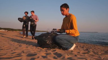Dünya günü. Gönüllü eylemciler, sahil bölgesindeki çöpleri topluyor. Kadın, okyanus kıyısındaki çöp poşetine plastik şişe koyuyor. Çevre koruma kıyı bölgesi temizliği
