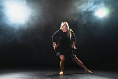 Siyah arka planda dumanla poz veren çağdaş kadın dansçı. Yetişkin kadın sanatçı ifade ile dans ediyor. Modern balerin karanlıkta dans ediyor. Tutku karanlıkta dans eder. Esneklik dengesi