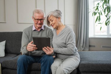Tabletli yaşlı çift internetten video izliyor. Karı koca evde vakit geçiriyor. Mutlu modern olgun yaşlı çift aile eğlencesi için tablet kullanıyor. Yaşlılar modern teknoloji