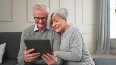 Tabletli yaşlı çift internetten video izliyor. Karı koca evde vakit geçiriyor. Mutlu modern olgun yaşlı çift aile eğlencesi için tablet kullanıyor. Yaşlılar modern teknoloji