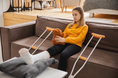 Kanepede oturan kırık bacaklı genç kız. Üzgün küçük çocuk, kırık bacağını iyileştirmek için oyuncak ayısına sarılıyor. Alçı ve koltuk değneği kullanan bir kız. Kaza sonucu oluşan kırık, ayak bandajı.