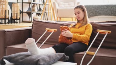 Kanepede oturan kırık bacaklı genç kız. Üzgün küçük çocuk, kırık bacağını iyileştirmek için oyuncak ayısına sarılıyor. Alçı ve koltuk değneği kullanan bir kız. Kaza sonucu oluşan kırık, ayak bandajı.