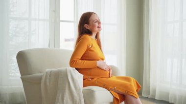 Hamilelik anneliği insanların gelecek beklentisi. Koca göbekli hamile bir kadın evdeki pencerenin yanındaki sandalyeye oturuyor. Karnına sarılan ve hamileliğin tadını çıkaran bir kız. Annelik şefkati ebeveynlik yeni bir hayat