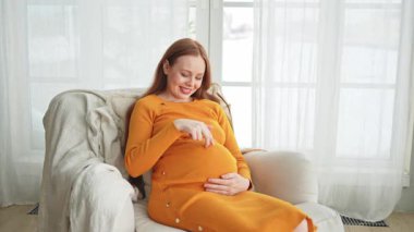 Hamilelik anneliği insanların gelecek beklentisi. Koca göbekli hamile bir kadın evdeki pencerenin yanındaki sandalyeye oturuyor. Karnına sarılan ve hamileliğin tadını çıkaran bir kız. Annelik şefkati ebeveynlik yeni bir hayat