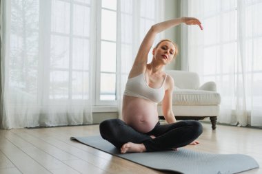 Hamilelik yoga antrenmanı. Hamile kadın evde yoga yapıyor. Hamile kız içeride yoga minderiyle spor yapıyor. Koca göbekli bir kadın Asya antrenmanı yapıyor.