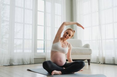 Hamilelik yoga antrenmanı. Hamile kadın evde yoga yapıyor. Hamile kız içeride yoga minderiyle spor yapıyor. Koca göbekli bir kadın Asya antrenmanı yapıyor.