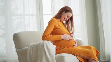 Hamilelik anneliği insanların gelecek beklentisi. Koca göbekli hamile bir kadın evdeki pencerenin yanındaki sandalyeye oturuyor. Karnına sarılan ve hamileliğin tadını çıkaran bir kız. Annelik şefkati ebeveynlik yeni bir hayat