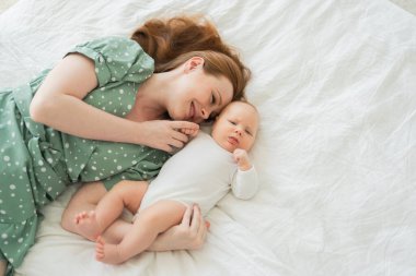 Evde mutlu bir aile. Anne, küçük bebekle yatakta oynuyor. Anne bebek birlikte oynayıp rahatlıyor. Anne bebeğe sevgiyle sarılır. Bebeği emziren anne oturma odasında dinleniyor.