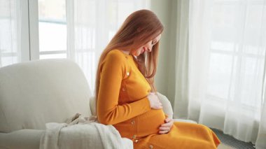 Hamilelik anneliği insanların gelecek beklentisi. Koca göbekli hamile bir kadın evdeki pencerenin yanındaki sandalyeye oturuyor. Karnına sarılan ve hamileliğin tadını çıkaran bir kız. Annelik şefkati ebeveynlik yeni bir hayat