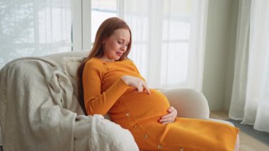 Hamilelik anneliği insanların gelecek beklentisi. Koca göbekli hamile bir kadın evdeki pencerenin yanındaki sandalyeye oturuyor. Karnına sarılan ve hamileliğin tadını çıkaran bir kız. Annelik şefkati ebeveynlik yeni bir hayat