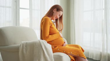 Hamilelik anneliği insanların gelecek beklentisi. Koca göbekli hamile bir kadın evdeki pencerenin yanındaki sandalyeye oturuyor. Karnına sarılan ve hamileliğin tadını çıkaran bir kız. Annelik şefkati ebeveynlik yeni bir hayat