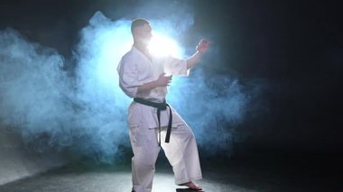 Siyah kuşaklı karate dövüş sanatları için beyaz kimonolu güçlü adam siyah arka plandaki dövüş eğitimine hazırlanıyor. Antrenman hareketleri yumruklar gölge dövüşü alıştırmaları antrenmana hazır.