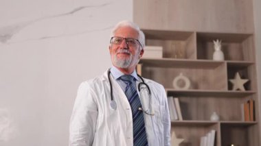 Gözlüklü yaşlı doktor hastane ya da doktor muayenehanesinde kameraya gülümsüyor. Pratisyen hekim, klinikte profesyonel sağlık uzmanı. Sağlık hizmetleri kavramı