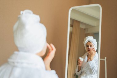 Yetişkin, yaşlı bir kadın aynaya bakıyor ve günlük güzellik kremi sürüyor. Cildinde beyaz maske kremi olan yaşlı bir kadın cilt bakımı görüyor.